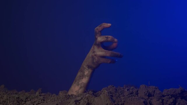 Scary dirty halloween zombie hand raising up from grave ground points finger at side space, isolated on blue background. Mystery halloween concept.