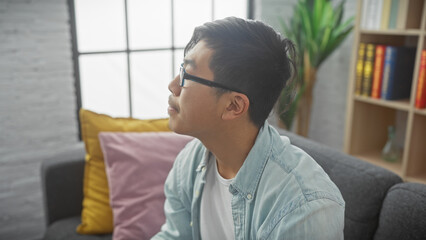 Pensive young asian man with glasses looking away while sitting on a sofa in a cozy living room.