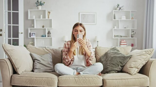 Young woman in casual clothes is sitting on a sofa in a living room, holding a smartphone and apparently interacting with it