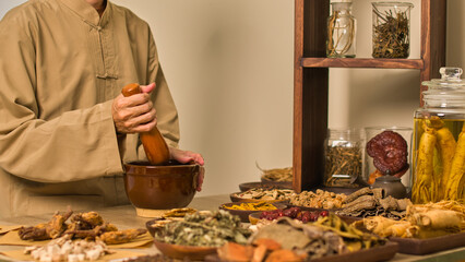 A traditional herbalist wearing a brown robe gathers everyone around a table where they peel and crush herbs using a mortar and pestle before arranging them on wooden trays to soak in glass jars.