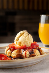 A hand pours creamy sauce over a stacked dessert featuring waffles, vanilla ice cream, and fresh strawberries, alongside a refreshing glass of orange juice.