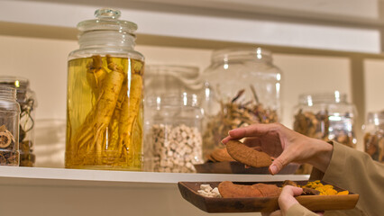 A photo of a traditional Asian medicinal chamber features the scientist delicately places and arranges a pile of reishi mushrooms on a plate on a shelf, adjacent to jars and bottles of other herbs.