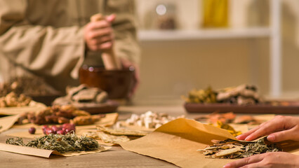 One herbalist in traditional brown robes is pounding dried herbs, while another picks up a package...