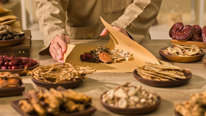 A doctor meticulously wrapping dried herbs in brown paper and surrounding the table with other...