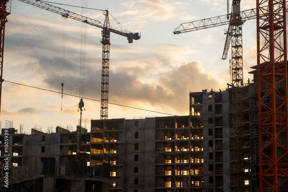 Poster Multiple cranes work efficiently at a construction site as the sun sets, illuminating the building's framework and reflecting on workers below