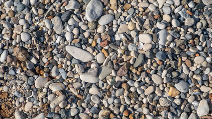 Smooth pebbles of various colors and sizes cover the sandy beach, illuminated by sunlight on a clear day