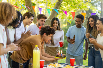 A multiracial group of friends celebrates a birthday outdoors with cake and colorful decorations.