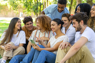 A multiracial group of friends sits on the grass, smiling and enjoying a moment together while looking at a smartphone.