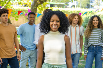 A smiling young woman stands in focus at the front while her friends gather behind her outdoors.