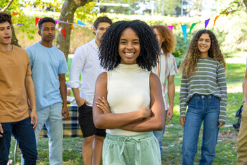 A confident afro american young woman with arms crossed smiles in the foreground, surrounded by friends outdoors.