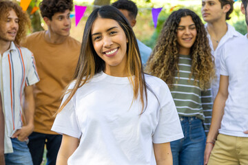 A smiling young latin woman stands in the foreground while a group of friends gathers behind her outdoors.