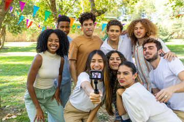 A multicultural group of smiling friends gathers closely for a selfie with a selfie stick at an outdoor event, surrounded by colorful decorations.
