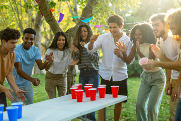 A group of friends plays a game with red and blue cups on a table, cheering and having fun during an outdoor gathering.