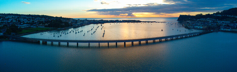 Fototapeta premium Teignmouth sunrise aerial panoramic picture. Dark landscape with a warm glowing sunrise. South Devon tourism. Shaldon bridge and port. 