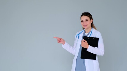 Happy Caucasian female young doctor or nurse pointing finger left to copy space on gray background. Concept of a medical advertisement.