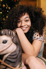 Close-up portrait of young African woman in elegant dress near Christmas tree at home. Charming black girl near vintage toy.