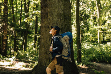Fototapeta premium Side view of a senior male with a tourism backpack standing in a forest near a big tree. Mature tourist enjoying his forest walk.