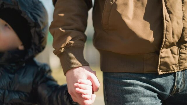 A close up of an adult and a child walking hand in hand, the child raises his ight hand up, the adult is dressed in a brown jacket and jeans, while the child is wearing a shiny black jacket