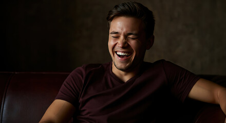Fototapeta premium Smiling young man laughing on a leather couch. Indoor portrait with dark background
