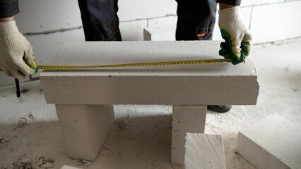 A worker measures the length of a building block for an internal partition. A builder uses a tape measure at a construction site. During the process of insulating walls at a construction site to cut