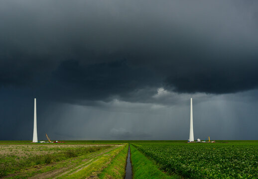 Dark clouds over two wind turbines under construction in a green agricultural field.