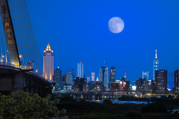 A golden autumn moon hangs over the Taipei skyline, casting a warm glow on the city. The scene captures the tranquility of an autumn evening. Taipei City, Taiwan.