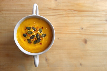 Bowl of creamy pumpkin and potato soup, with pumpkin seeds and chilli flakes toppings. Top view, wooden background.
