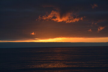 Fototapeta premium A stunning sunset over the ocean with dramatic dark clouds and a vivid orange and red horizon, casting warm reflections on the calm water