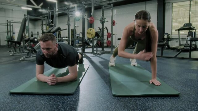 Woman performing plank with shoulder taps while man doing elbow plank hip dips on mats in gym during core and stability workout session
