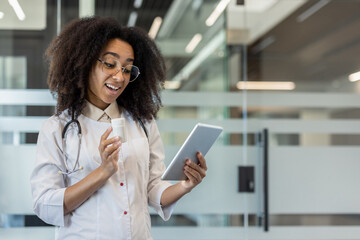 Fototapeta premium Photo of a young African-American female doctor in a hospital room, praying on a video call on a tablet, holding a jar of pills in her hand and showing the camera