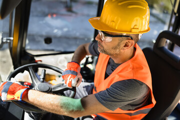 Construction worker operating heavy machinery at a job site during daytime wearing safety gear and...
