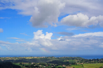 Rayo de arco iris en cielo nublado sobre litoral rural de Asturias