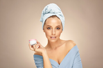 Young caucasian woman after shower in spa bathrobe and towel on head, presenting face cream in white jar on palm applying mask for flawless skin on beige isolated background.