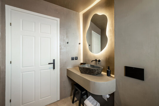Modern bathroom interior design with cave-like aesthetic, featuring soft beige tones. Focal point includes unique stone-like sink with natural finish, unusually shaped mirror with stylish backlighting