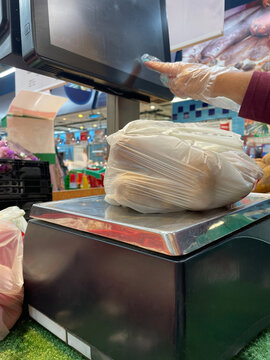 A gloved hand operates a digital screen while weighing a bag of potatoes at a market produce scale station.