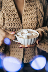 Woman in a knitted sweater holding a cup with coffee, hot chocolate, or cocoa drink with marshmallow. Close-up view