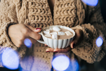 Woman in a knitted sweater holding a cup with coffee, hot chocolate, or cocoa drink with marshmallow. Close-up view