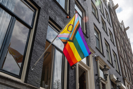 Progress Pride flag with intersex symbol displayed on a building facade in an urban street.