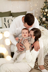 a young family with a small child is enjoying the New year at the Christmas tree on the bed, smiling, kissing and hugging congratulating each other celebrates the holiday