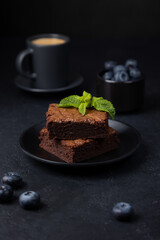 Chocolate brownie cake with mint, blueberries and espresso. Traditional American dessert. Popular cake with dark chocolate on a black background. Close-up, selective focus.