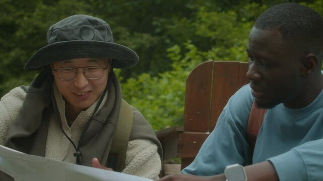 Waist up of two young ethnically diverse male tourists looking at paper map and planning route of their hiking trip while resting in camping area in woods