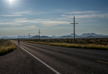 Fototapeta premium A lone cyclist riding on a straight section of paved road long a desert scene with power line and poles stretching to the horizon, Davis Mountain, Texas