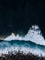 Top view from drone of rocks and cube stones on the beach with big waves in stormy ocean on sunset - vertical photo. Blue Atlantic ocean with azure water from above - travel concept.