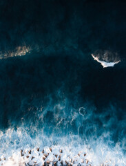 Top view from drone of rocks and cube stones on the beach with big waves in stormy ocean on sunset - vertical photo. Blue Atlantic ocean with azure water from above - travel concept.