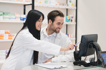 Fototapeta premium Young asian pharmacist pointing showing male colleague medication prices, recipes on computer screen in white medical coats at cash point desk in drugstore