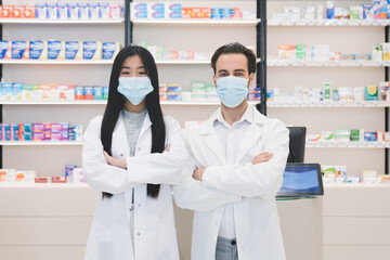 Two confident successful young pharmacists druggists in white medical coats in protective face masks against coronavirus standing with arms crossed at the cash point desk in pharmacy drugstore