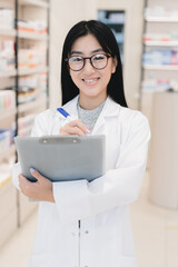 Vertical portrait of asian female young pharmacist druggist in white medical coat writing on clipboard side effects, active substance, prescriptions standing at pharmacy drugstore