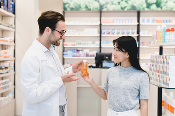 Fototapeta premium Consultation. Caucasian male young pharmacist druggist advising medicines, pills, drugs, painkillers, vitamins to a female asian customer client, buyer in drugstore