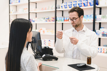 Male caucasian handsome pharmacist druggist reading medical prescription for medications, drugs, painkillers, antibiotics, pills of female asian client customer buyer in pharmacy