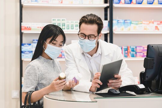 Male pharmacist druggist in protective face mask against coronavirus advising female customer client buyer remedy, medicines, pills, antibiotics against Covid19 using digital tablet in pharmacy